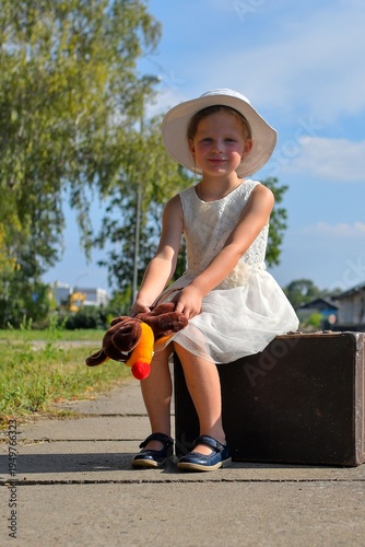 A little girl on a train platform. A cute girl at a train station. A little girl holds a stuffed toy and sits on an old suitcase. The concept of travel, holidays, and a carefree childhood. Retro and