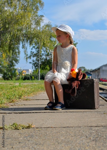 A little girl on a train platform. A cute girl at a train station. A little girl holds a stuffed toy and sits on an old suitcase. The concept of travel, holidays, and a carefree childhood. Retro and