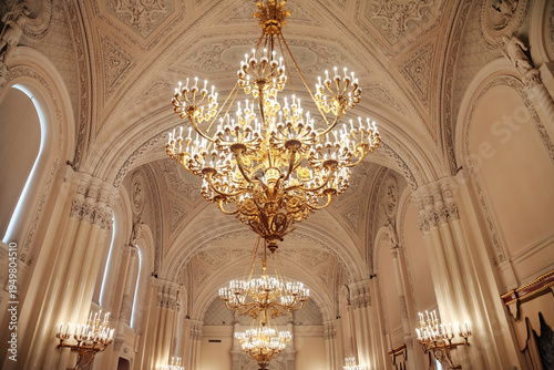Multiple magnificent golden chandeliers hang from an intricately sculpted ceiling in a luxurious, historical hall.