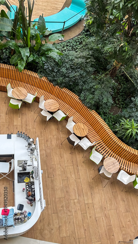 Modern cafe interior with wooden tables, chairs, curved bench, lush green plants, and a bar