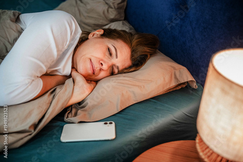 Canvas Print Tired middle aged woman sleeping in bed after using smartphone.