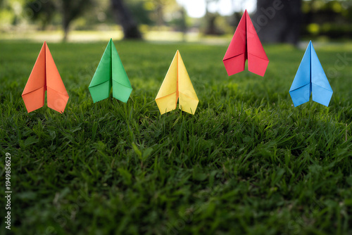 Five colorful paper airplanes standing upright on green grass in a park setting with natural background