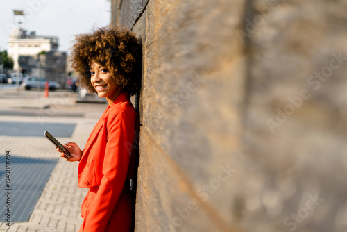Portrait of smiling young woman with cell phone wearing fashionable red pantsuit leaning against wall