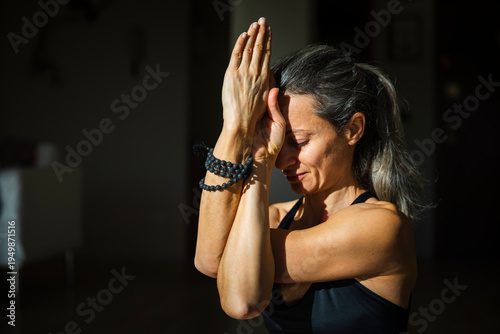 Mature woman practicing yoga with twisting arms at home on sunny day