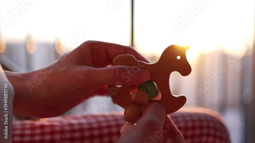 Close-up view of a woman's hands holding a charming wooden rocking horse teether, gently touching its smooth surface as the warm sunlight streams through the window at sunset