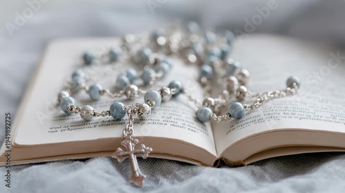 A close-up of a rosary made of blue beads resting on an open book. The book has a soft cover and is placed on a textured fabric surface.