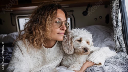 A young Hispanic woman with curly hair sits in a van, gazing out the window. A small white dog rests beside her, both looking relaxed and contemplative.