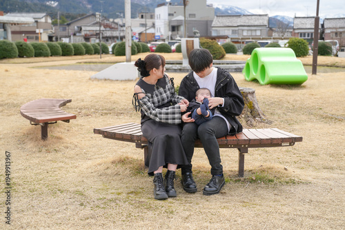 Cozy family moment on a park bench. Mother and father hold their baby gently. Soft smiles reflect quiet contentment. Background shows green sculpture and distant hills. Ideal for parenting, family