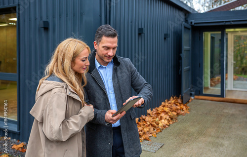 Architect explaining design concepts of modern modular house built from repurposed shipping containers with solar panels in digital tablet to a satisfied client, with autumn leaves on the ground