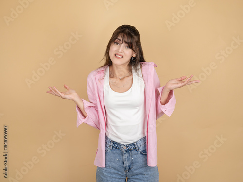 Confused young woman shrugging shoulders with open palms, wearing pink shirt and denim, standing against beige studio background with wide copy space. Uncertainty and doubt concept.