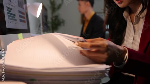 Businesswoman's hands sorting through a large stack of papers at her desk while working late at night, analyzing data charts on a computer.