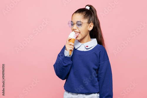 Fotografie Little kid girl 8-9 years old she wear blue shirt sunglasses have fun eating ice-cream isolated on pastel light pink color background child studio portrait