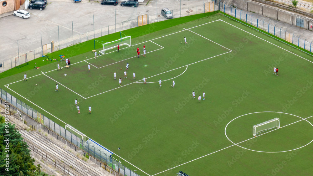 Obraz premium Aerial close-up of a vibrant green synthetic soccer pitch where two teams are engaged in a match. The field features white markings, two goals, and several players scattered across the turf.