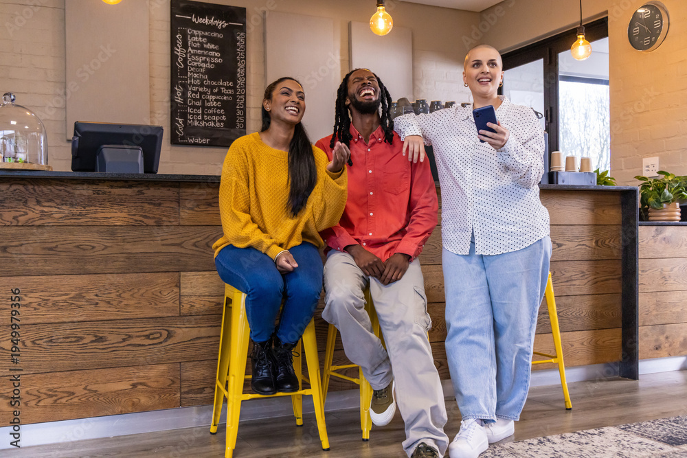 Fototapeta premium Diverse friends laughing and chatting at cafe counter, holding smartphone with two yellow stools