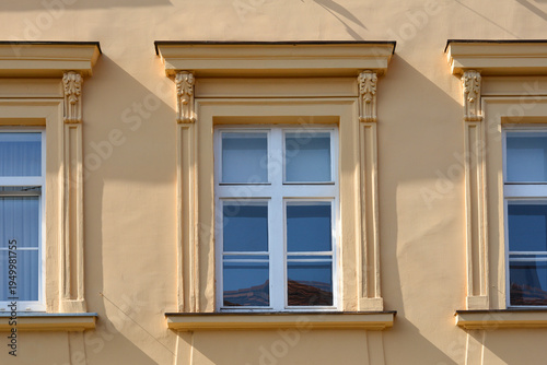 Three white windows are set into a yellow building facade featuring elegant architectural moldings and decorative ornaments above each frame