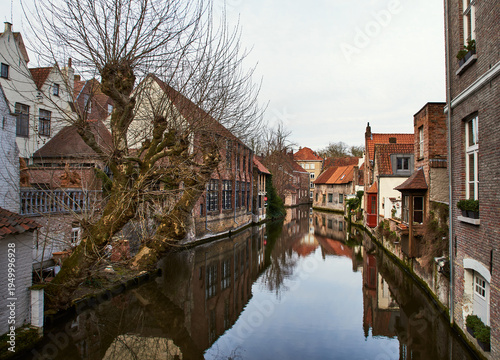 Serene canal in historic Belgian town Bruges. Medieval brick houses with gables reflect in calm water conveys concepts of heritage architecture, seasonal melancholy and urban waterway symmetry