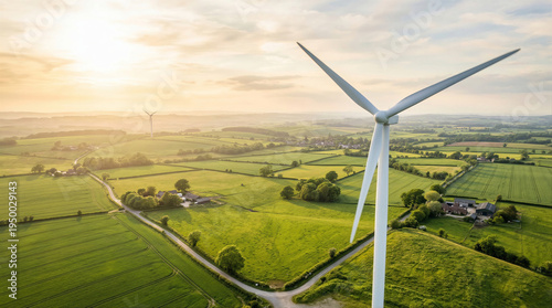 Aerial view of a white wind turbine in a lush green countryside at sunrise with rolling fields and farmhouses