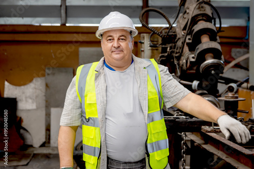 Portrait of happy engineer supervisor man with safety vest and white helmet stands at manufacturing plant metal factory. Senior industrial foreman worker working and maintaining machine at factory.