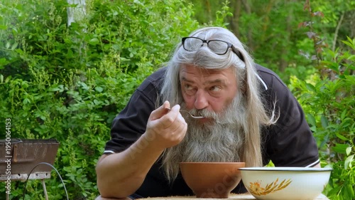 Close-up portrait of a senior bearded man tasting homemade soup with a spoon in a summer garden.
