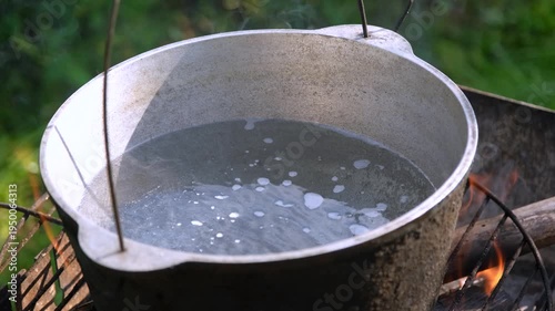 Close-up of water boiling with steam in a metal cauldron over an open campfire flame outdoors.