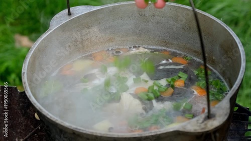 Close-up of chopped green onions being added to fish soup boiling in a cauldron outdoors.