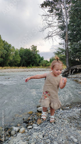 A little girl plays on the bank of a mountain river on a cloudy evening. She runs and stands by the water. An atmosphere of calm, natural freedom, the joy of childhood, and closeness to nature.