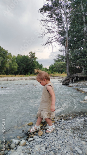 A little girl plays on the bank of a mountain river on a cloudy evening. She runs and stands by the water. An atmosphere of calm, natural freedom, the joy of childhood, and closeness to nature.