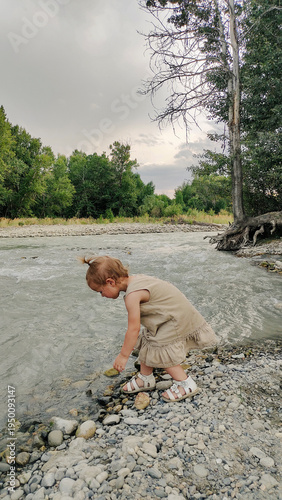 A little girl plays on the bank of a mountain river on a cloudy evening. She runs and stands by the water. An atmosphere of calm, natural freedom, the joy of childhood, and closeness to nature.
