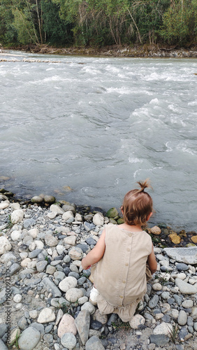 A little girl plays on the bank of a mountain river on a cloudy evening. She runs and stands by the water. An atmosphere of calm, natural freedom, the joy of childhood, and closeness to nature.