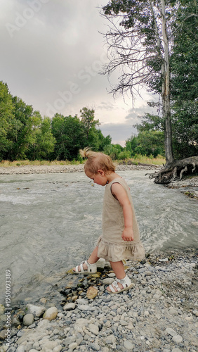 A little girl plays on the bank of a mountain river on a cloudy evening. She runs and stands by the water. An atmosphere of calm, natural freedom, the joy of childhood, and closeness to nature.