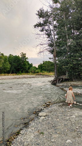 A little girl plays on the bank of a mountain river on a cloudy evening. She runs and stands by the water. An atmosphere of calm, natural freedom, the joy of childhood, and closeness to nature.