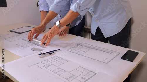 Two architects drawing a blueprint, collaborating on a house project. An engineer and a draftsman planning building construction using technical drawing tools on a desk