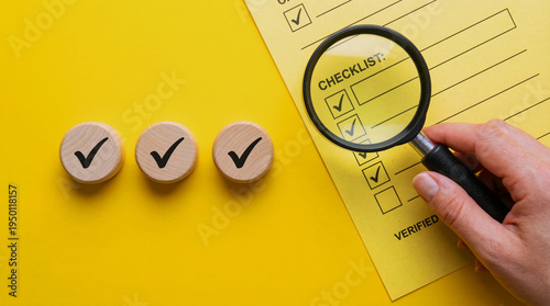 A hand holding a magnifying glass over a checklist and three wooden checkmarks on a yellow background