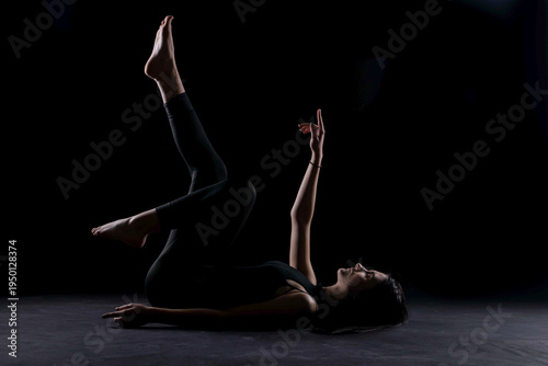 Fit young woman in black sportswear lies on the floor performing a core or Pilates-style leg raise exercise, one leg lifted and arms extended upward in dramatic studio lighting dark background