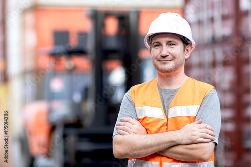 Portrait of happy engineer supervisor man with safety vest and white helmet standing with arms crossed during inspects containers. Foreman worker working at logistic shipping cargo container yard.