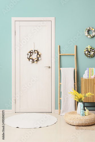 Interior of festive room with commode, ladder, ottoman, flowers and Easter wreaths hanging on blue wall