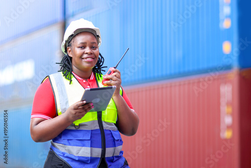 African woman engineer wearing safety vest and helmet, holding worky talky and tablet while inspecting the containers. Female foreman worker working at logistic shipping cargo container yard.