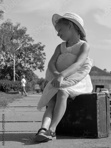 Young girl in a hat sits on an old suitcase, looking down with a pillow