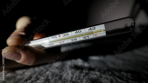 Person hand holds a glass mercury thermometer showing a dangerously high temperature over 39 degrees Celsius. A close-up in a dark room highlights the urgent medical reading.
