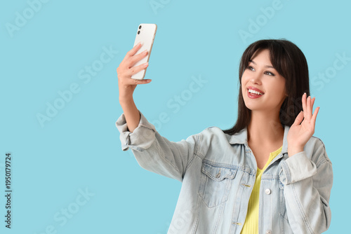 Smiling young woman with mobile phone taking selfie on blue background