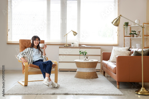 Konstfotografi Young woman with headphones and tea cup sitting in leather armchair at home