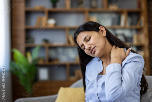 Young woman grimacing indoors, rubbing sore neck and shoulder from fatigue and poor posture, showing stress, muscle tension and discomfort while sitting at home