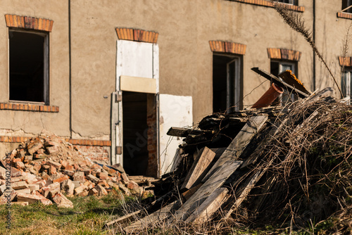 Rubble and wooden debris gather in front of a rundown building. Two windows show signs of neglect. The sunlight highlights the piles of materials on the grass