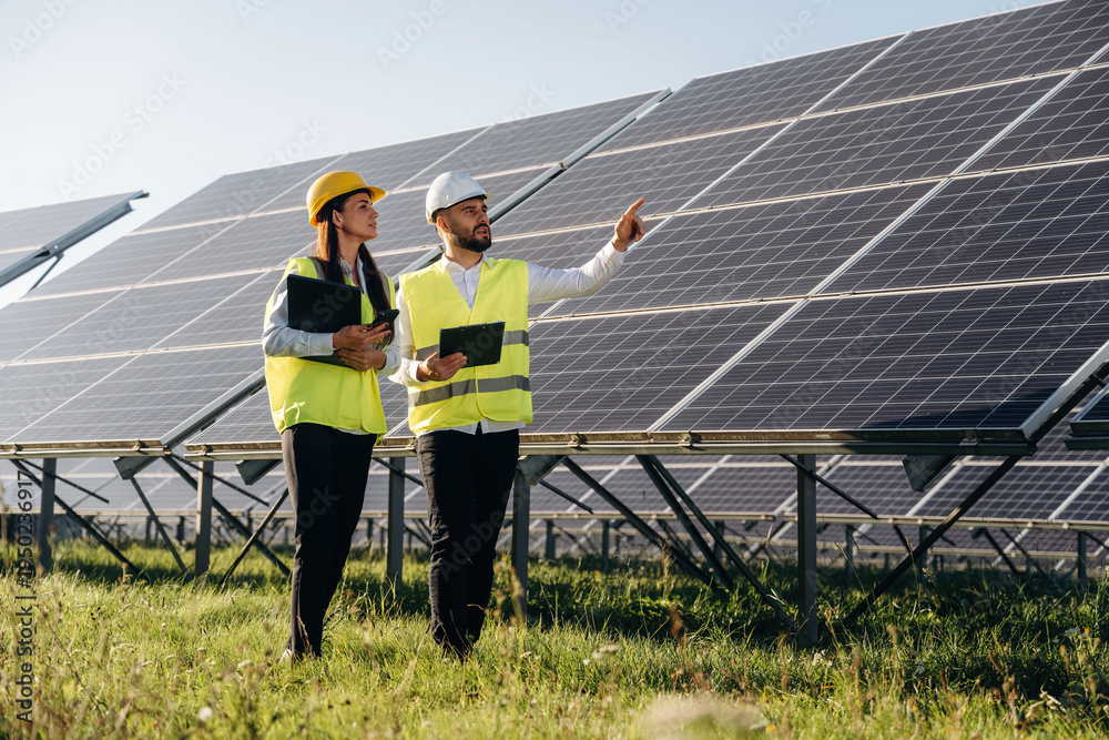 Obraz premium Explaining the plan and showing are to the woman. Two solar engineers standing together at a photovoltaic field, in bright safety vests