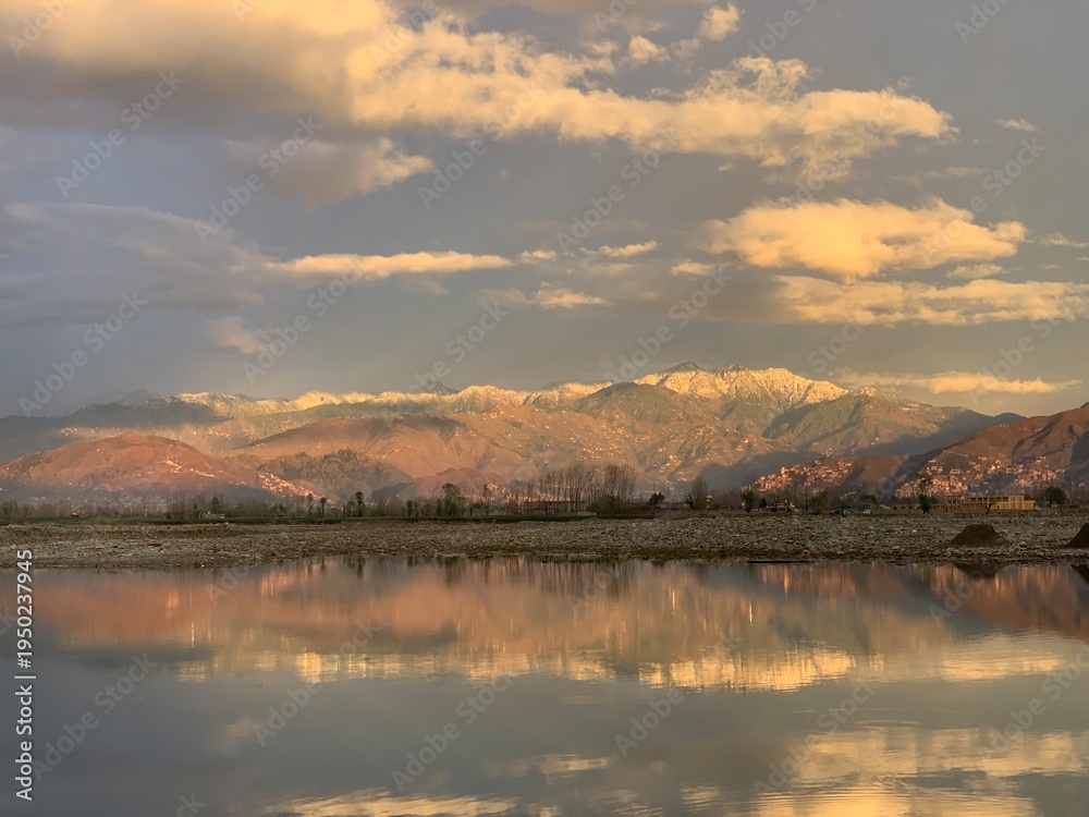 Obraz premium A scenic wide-angle landscape featuring a calm river in the foreground with a crisp mirror reflection of the surrounding environment. In the background, majestic mountains are bathed in the warm.
