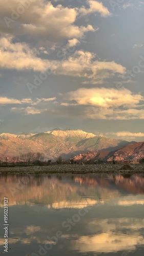 A scenic wide-angle landscape featuring a calm river in the foreground with a crisp mirror reflection of the surrounding environment. In the background, majestic mountains are bathed in the warm.