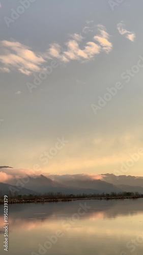 A scenic wide-angle landscape featuring a calm river in the foreground with a crisp mirror reflection of the surrounding environment. In the background, majestic mountains are bathed in the warm.