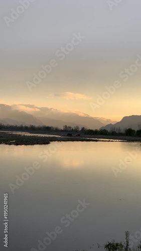 A scenic wide-angle landscape featuring a calm river in the foreground with a crisp mirror reflection of the surrounding environment. In the background, majestic mountains are bathed in the warm.