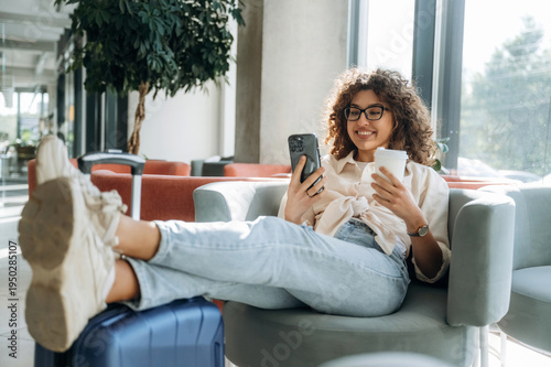 Relaxing on chair and using phone. Beautiful woman is in airport hall with suitcase