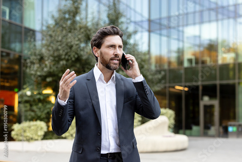Businessman in a suit standing in front of a modern glass office building, speaking on a smartphone and gesturing with his hand during a focused outdoor conversation
